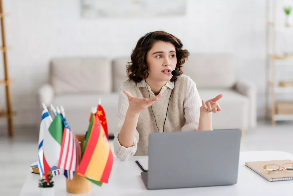brunette-language-teacher-headset-gesturing-near-laptop-blurred-international-flags-home