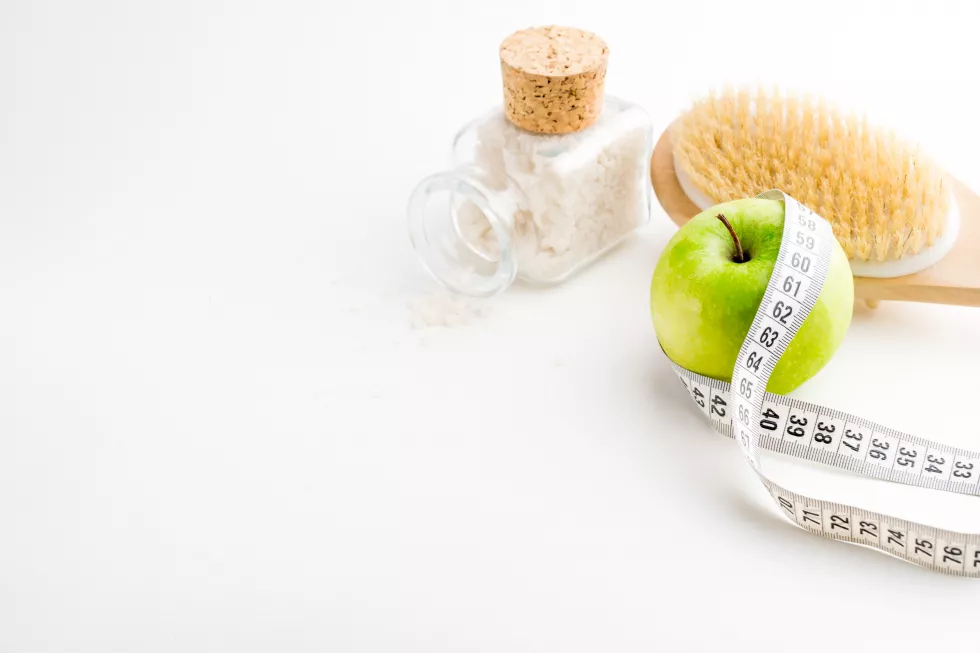 dry-massage-brush-with-tape-measure-single-green-apple-glass-bottle-with-sea-salt-white-desk-health-diet