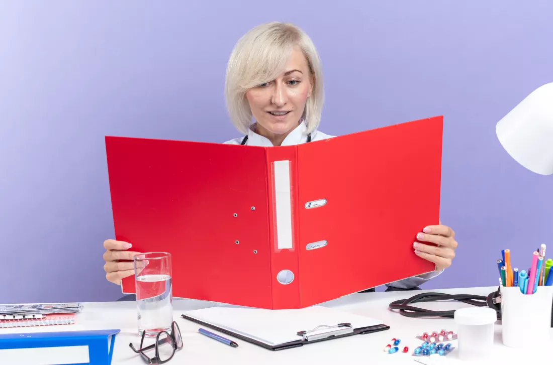 pleased-adult-slavic-female-doctor-medical-robe-with-stethoscope-sitting-desk-with-office-tools-holding-looking-file-folder-isolated-purple-wall-with-copy-space