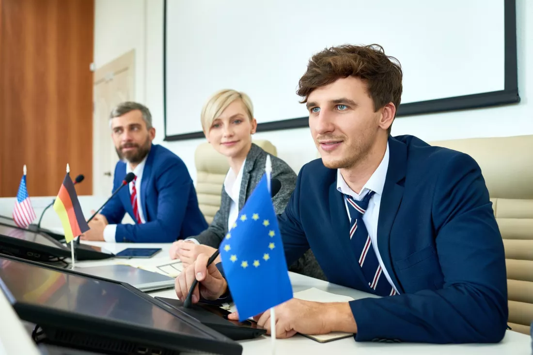 Młodzi delegaci w garniturach przy stole obrad z flagami państw i Unii Europejskiej