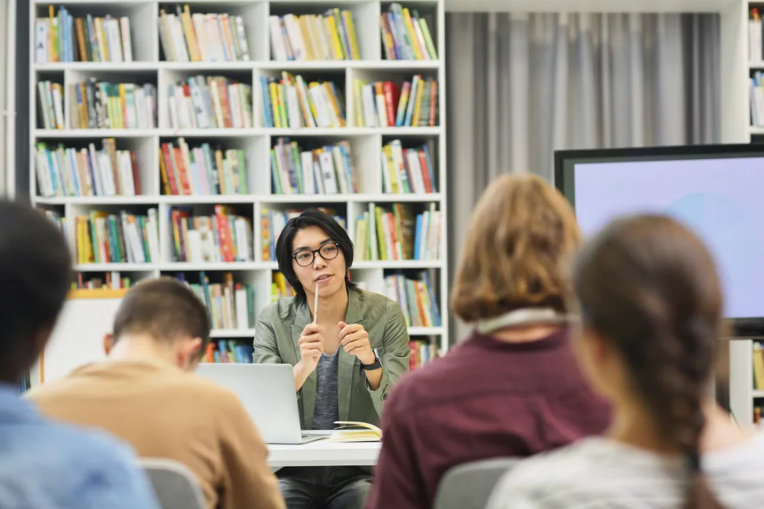 Studenci podczas zajęć seminaryjnych w bibliotece, prowadząca tłumaczy coś grupie.