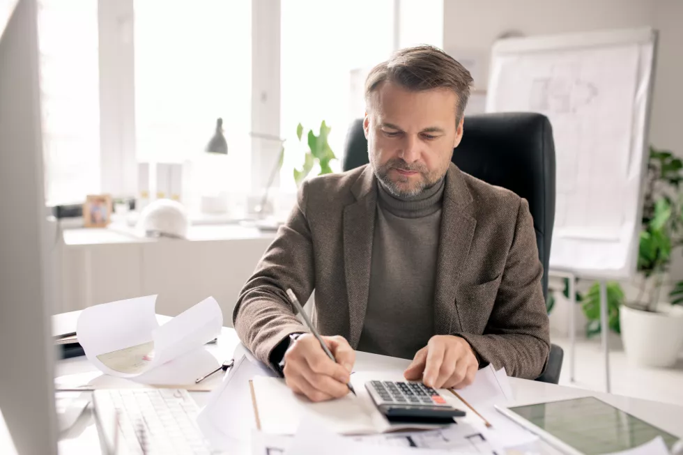 serious-mature-engineer-accountant-with-pen-calculator-making-notes-notebook-while-sitting-by-desk-office