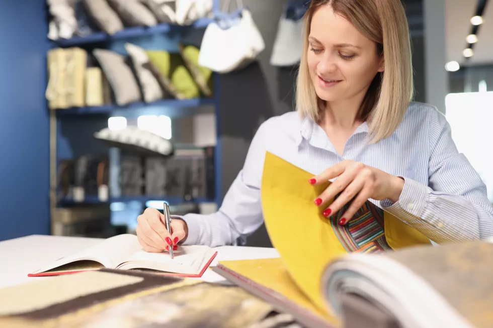 woman-writing-with-pen-notebook-leafing-through-catalog-with-fabrics-sewing-curtains