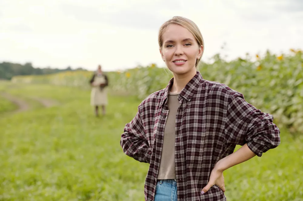 young-blond-smiling-female-farmer-workwear-looking-you-while-standing-front-camera-using-digital-tablet-against-green-field