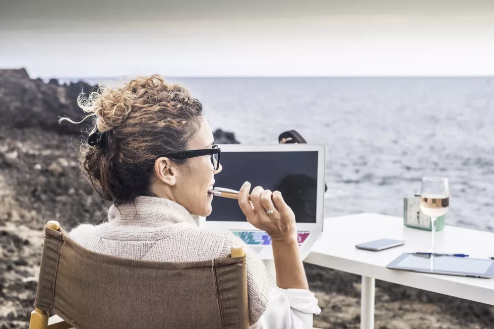 rear-view-mature-woman-using-laptop-while-sitting-chair-beach-against-sky-sunset