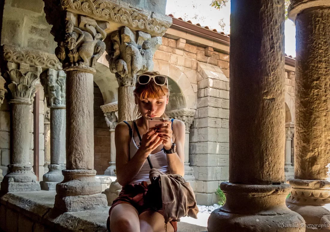 woman-using-mobile-phone-while-sitting-retaining-wall-historic-building-corridor